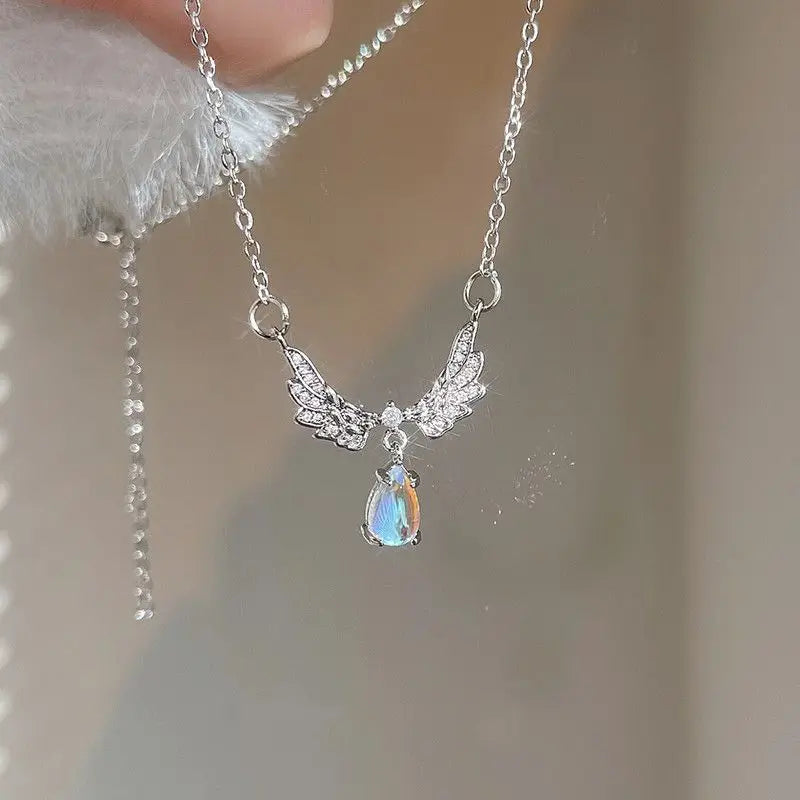 A silver-colored angel wings pendant necklace with a moonstone in the center, held between fingers.